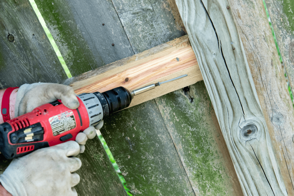 someone wearing white dirty gloves and using a red drill to replace a section of a wooden fence